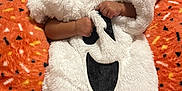 V'Niyah is registered to the contest to win money with this photo: baby, child, costume, ghost, white, orange_blanket, cute, surprised, hooded, fluffy, infant, patterned_blanket, lying_down, indoor, portrait, soft_texture, hands, face, expression, holiday