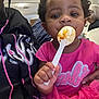 toddler, child, pink_dress, spoon, food, indoor, person, face, hand, curly_hair, seated, adult_hand, table, casual, closeup, expression, cute, eating, clothing, people