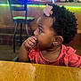 toddler, child, girl, hair_accessory, flower_clip, curly_hair, pink_clothing, hand_on_mouth, table, wood, indoor, bar, chair, colorful_lights, background, side_view, portrait, young_child, seated, casual