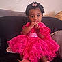 toddler, child, pink_dress, curly_hair, hair_clip, shoes, couch, indoor, portrait, cute, baby, sitting, hand_in_mouth, home, furniture, soft_lighting, curiosity, young_child, casual, cozy