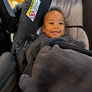 Ahlegend is registered to the contest to win money with this photo: baby, blanket, bundled, car_seat, child, cozy, dark_clothing, eyes, face, hair, happy, indoor, infant, photograph, portrait, safety_label, sitting, smiling, travel, vehicle