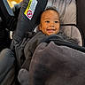baby, blanket, bundled, car_seat, child, cozy, dark_clothing, eyes, face, hair, happy, indoor, infant, photograph, portrait, safety_label, sitting, smiling, travel, vehicle