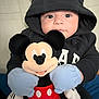 baby, infant, mickey_mouse, plush_toy, stuffed_animal, hoodie, mouse_ears, mittens, blue_mittens, black_hoodie, red_shorts, yellow_shoes, big_eyes, portrait, indoor, blanket, cute, smiling, toy, person