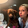 beauty_products, brown_dog, casual_clothing, cosmetics, dog, expression, face, fan, indoor, jewelry, lamp, lighting, nail_polish, pet, pink_glove, reflection, short_hair, table, woman, young_adult
