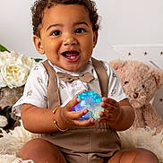 Dejai joined the competition — help win amazing prizes! baby_bracelet, bow_tie, child, close_up, curly_hair, cute, dinosaur_pattern, fluffy_blanket, glowing_orb, happy, holding_object, indoor, person, portrait, seated, smiling, suspenders, teddy_bear, toddler, white_flowers
