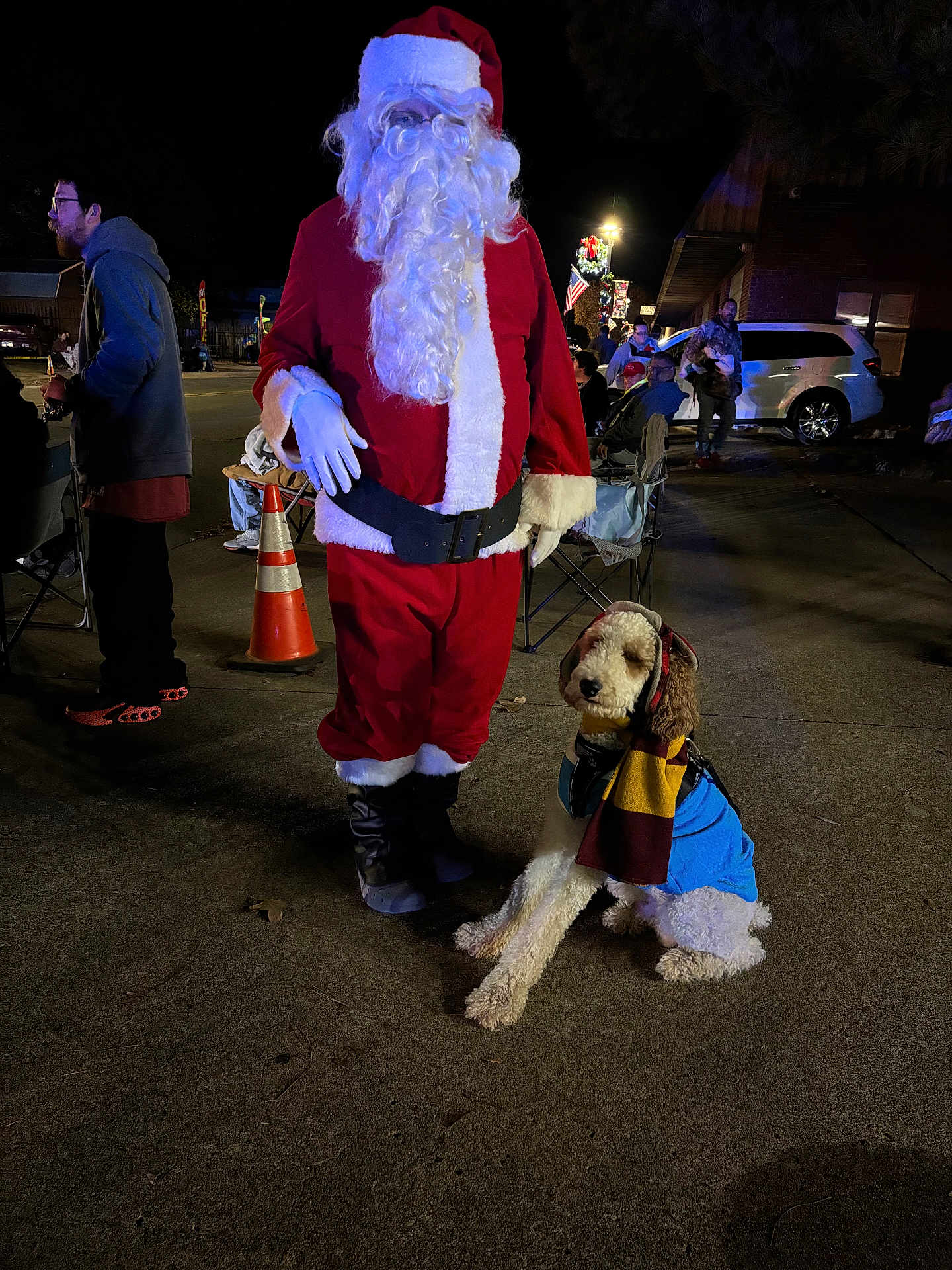 Puddles is registered to the contest to win money with this photo: person, dog, santa_claus, costume, scarf, night, outdoor, people, street, holiday, festive, lights, vehicle, chair, concrete, beard, glasses, hat, boots, jacket