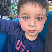 Julian is registered to the contest to win money with this photo: amusement_park, backpack, blue_shirt, boy, casual_clothing, child, closeup, daylight, expressionless, face, green_eyes, hair, outdoor, person, portrait, ride, rosy_cheeks, seat, usa, young_child