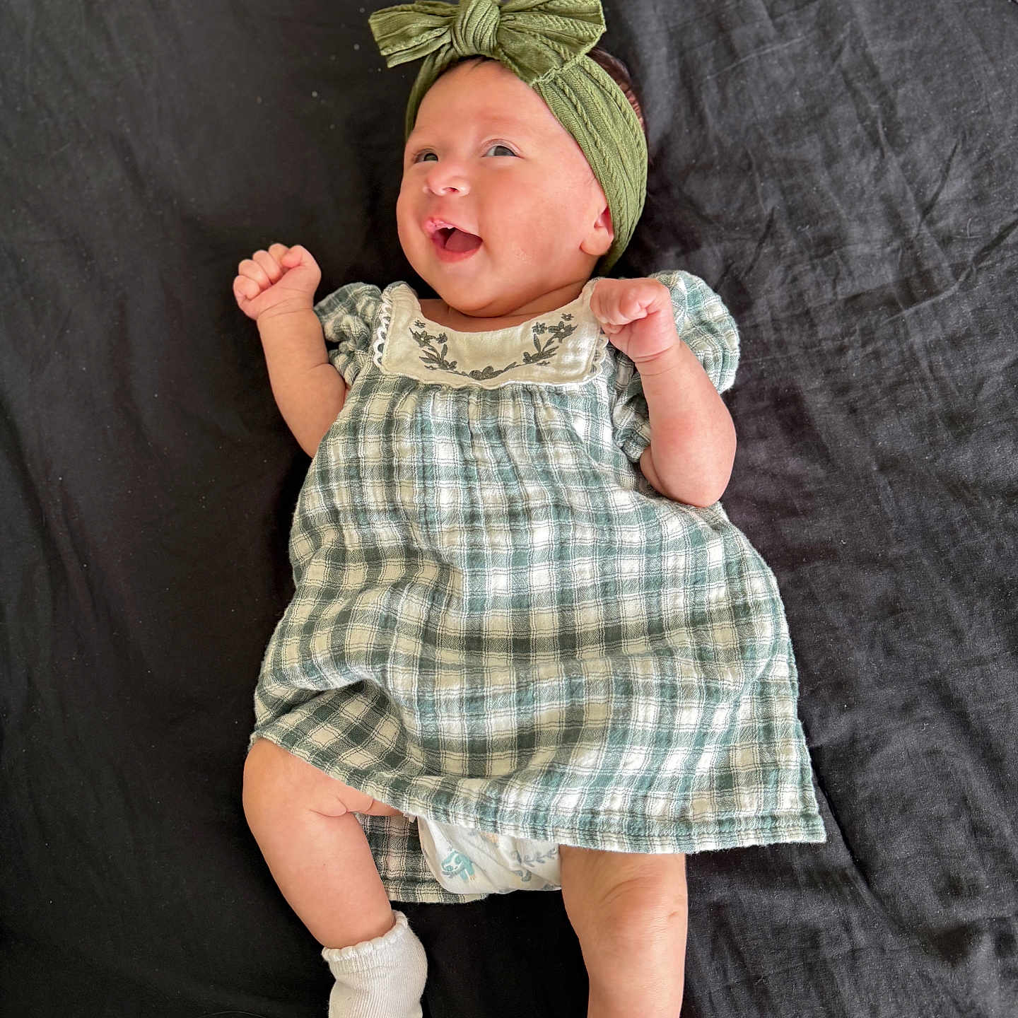 Kingsley is registered to the contest to win money with this photo: baby, infant, child, smiling, headband, green_bow, dress, plaid, socks, lying_down, blanket, happy, cute, small_hands, portrait, indoors, clothing, person, skin, toddler