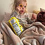 toddler, child, snack, food, blanket, couch, pillow, indoor, face, hand, blonde_hair, striped_shirt, curly_hair, playful, expression, cozy, wrapped, sitting, young_child, curious