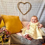 Piper joined the competition — help win amazing prizes! baby, child, dress, smile, beanbag, pillow, flower_arrangement, plush_toy, heart_decoration, brick_wall, indoor, flooring, yellow_pillow, happy, cute, sitting, person, decor, soft_furniture, cozy