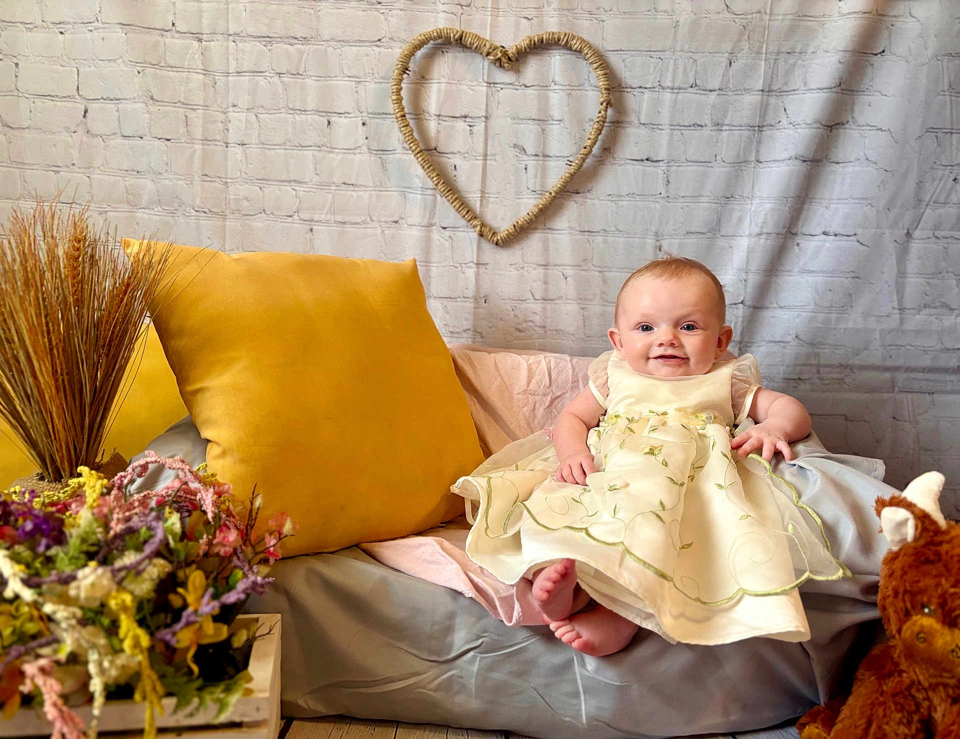 Piper joined the competition — help win amazing prizes! baby, child, dress, smile, beanbag, pillow, flower_arrangement, plush_toy, heart_decoration, brick_wall, indoor, flooring, yellow_pillow, happy, cute, sitting, person, decor, soft_furniture, cozy
