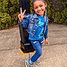 Navy is registered to the contest to win money with this photo: child, girl, smiling, peace_sign, denim_jacket, blue_shirt, ripped_jeans, sneakers, braided_hair, hair_bows, beaded_hair, outdoor, sidewalk, black_post, greenery, concrete, casual_clothing, happy, posing, portrait