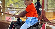 Navy joined the competition — help win amazing prizes! child, pony, saddle, orange_shirt, sneakers, indoor, horse_riding, cute, young_child, playful, background_people, wooden_structure, blurred_background, casual_clothing, jeans, pony_ride, red_blanket, happy, curious, afternoon_light