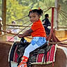 child, pony, saddle, orange_shirt, sneakers, indoor, horse_riding, cute, young_child, playful, background_people, wooden_structure, blurred_background, casual_clothing, jeans, pony_ride, red_blanket, happy, curious, afternoon_light