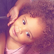 Ashanti participe au concours pour gagner de l'argent avec cette photo : child, toddler, curly_hair, pink_shirt, smile, face, eyes, skin, hand, laying_down, portrait, cute, indoors, comfort, happy, baby, person, young_child, head, expression