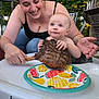 baby, woman, steak, high_chair, plate, outdoor, food, grilled_meat, smiling, happy, summer, nature, greenery, family, parent, child, curious, meal, picnic, table