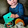 baby, blue_onesie, toy, colorful_toy, blanket, couch, patterned_couch, child, indoor, curious, face, hands, seated, portrait, infant, expression, soft_blanket, home, person, playtime
