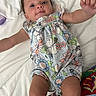 baby, infant, child, romper, floral_pattern, bed, white_bedding, cute, smiling, reaching_hand, person, young_child, lying_down, skin, face, blue_eyes, short_hair, indoors, colorful_clothing, playful