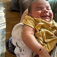 Jevonne is registered to the contest to win money with this photo: baby, smiling, yellow_clothing, arm, blanket, indoor, wooden_floor, person_holding, infant, happy, peaceful, sleepy, skin, human, closeup, portrait, newborn, cute, resting, warm_light