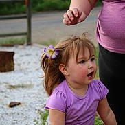 Deliah is registered to the contest to win money with this photo: toddler, child, flower, hair_clip, purple_shirt, adult, outdoor, gravel, road, nature, greenery, person, expression, casual_clothing, short_sleeves, daylight, side_view, cute, happy, playing