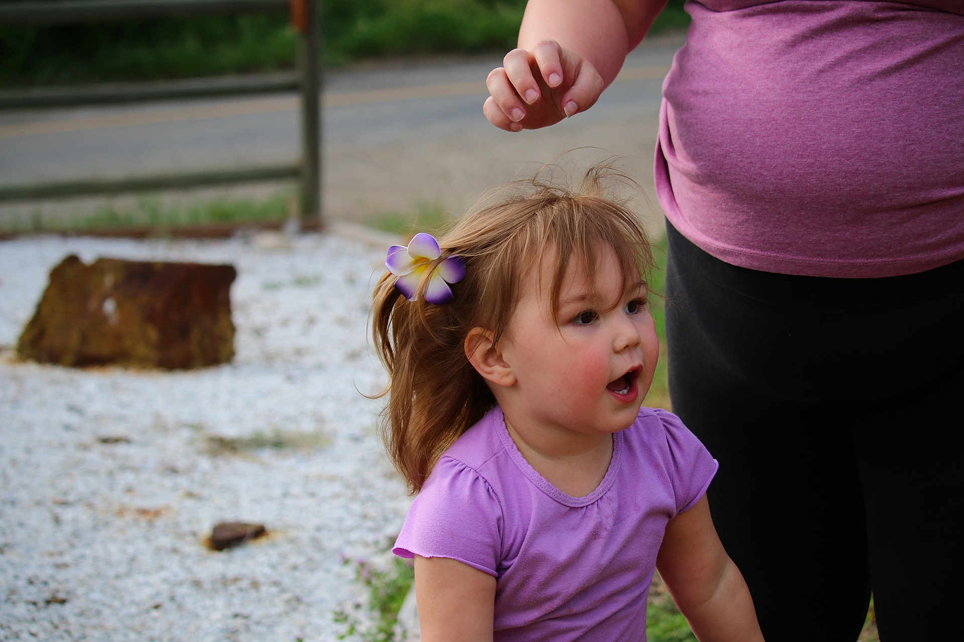 Deliah is registered to the contest to win money with this photo: toddler, child, flower, hair_clip, purple_shirt, adult, outdoor, gravel, road, nature, greenery, person, expression, casual_clothing, short_sleeves, daylight, side_view, cute, happy, playing