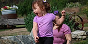 Deliah is registered to the contest to win money with this photo: child, toddler, purple_shirt, flower_hair, laughing, outdoor, adult, tattoo, grass, rocks, wheel, bench, greenery, garden, casual_clothing, happy, playful, person, nature, fun