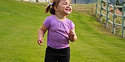 Deliah joined the competition — help win amazing prizes! child, toddler, running, grass, outdoor, flower, happy, smiling, purple_shirt, black_pants, sneakers, nature, mountains, fence, sky, cloudy, field, hair_accessory, playful, daytime