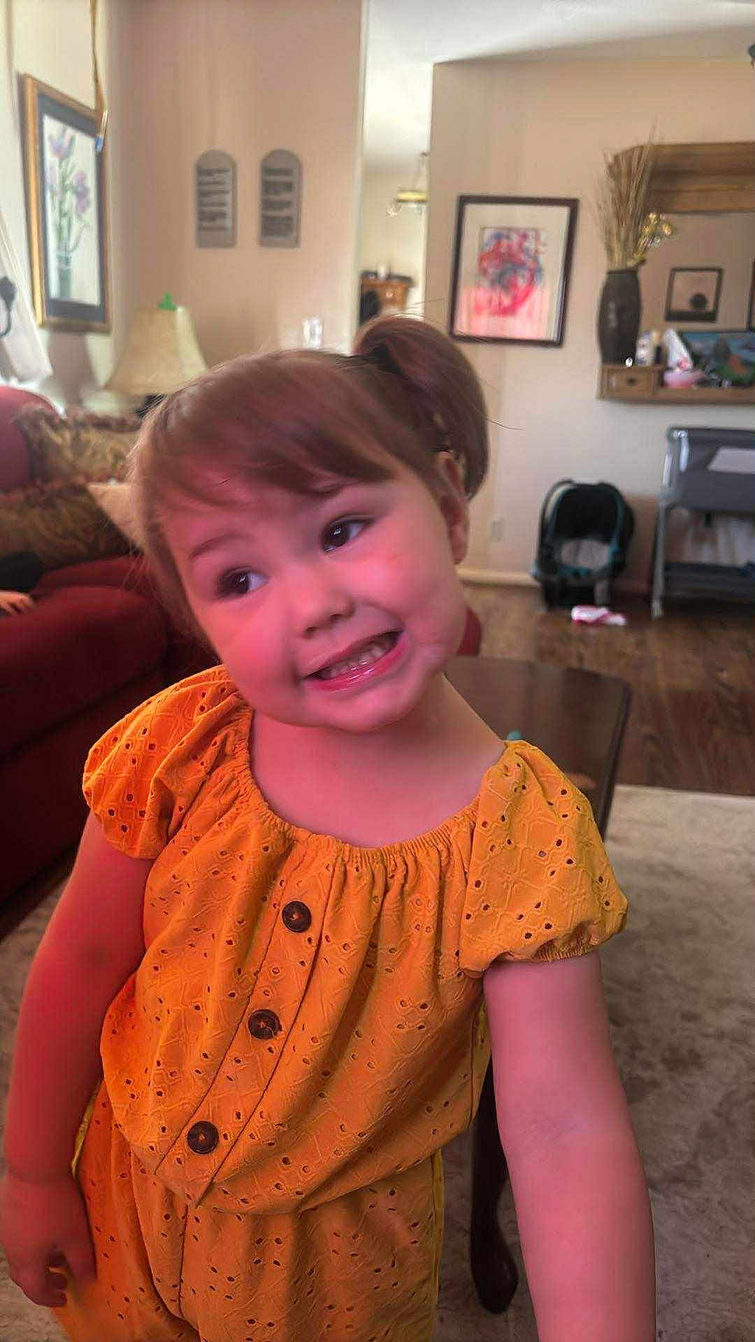 Deliah is registered to the contest to win money with this photo: child, girl, yellow_dress, smiling, playful, living_room, sofa, lamp, picture_frame, carpet, table, wood_floor, hair_tied, buttoned_dress, home_interior, furniture, cheerful, cute, person, portrait