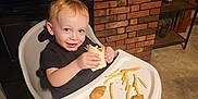 Colin is registered to the contest to win money with this photo: toddler, child, high_chair, food, chicken_nuggets, french_fries, wrap, kitchen, brick_wall, stove, smiling, blue_eyes, indoor, table, sitting, person, meal, happy, home, cute