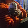 person, kitten, cat, white_cat, orange_sweater, glasses, holding, cuddling, indoor, window, warm_lighting, portrait, close_up, pet, animal, cute, cozy, hands, sitting, candid