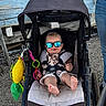 baby, stroller, sunglasses, child, toy, outdoor, person, hand, jeans, footwear, gravel, wood, blue, black, seat, clothing, sunshade, reflection, smile, casual