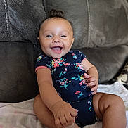 Naomi is registered to the contest to win money with this photo: baby, smiling, happy, infant, child, onesie, floral_pattern, couch, furniture, indoors, skin, hand, foot, hair, person, sitting, blanket, cute, portrait, expression