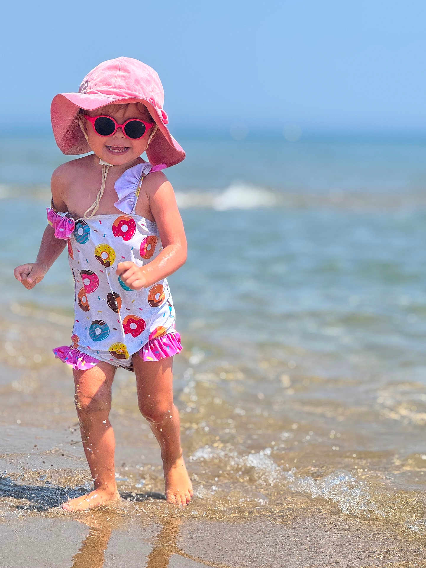 Lya a rejoint le concours — aidez-le/la à gagner de superbes lots ! toddler, child, beach, water, splashing, sunhat, sunglasses, swimsuit, donut_pattern, summer, outdoor, happy, running, sand, sea, sunny, barefoot, playful, holiday, sunshine