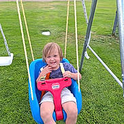 Travis joined the competition — help win amazing prizes! child, toddler, swing, playground, grass, smile, happy, outdoors, park, mountains, rope, metal_frame, blue_swing, red_buckle, little_tikes, shorts, tshirt, sandals, portrait, green_field