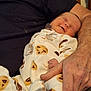 baby, infant, newborn, sleeping, smiling, pajamas, cookie_pattern, footed_pajamas, hand, adult_arm, holding, cozy, closeup, portrait, indoor, bedding, furniture, fabric, skin, affectionate