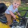 child, toddler, blue_eyes, blonde_hair, outdoor, toy_vehicle, steering_wheel, yellow, grass, dirt, firewood, curious, casual_clothing, sneakers, playful, sunlight, seat, hand, expression, person