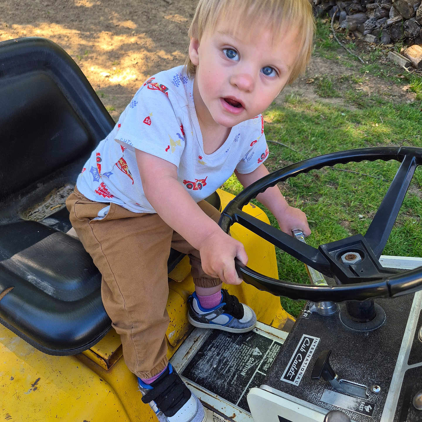 Jasper is registered to the contest to win money with this photo: child, toddler, blue_eyes, blonde_hair, outdoor, toy_vehicle, steering_wheel, yellow, grass, dirt, firewood, curious, casual_clothing, sneakers, playful, sunlight, seat, hand, expression, person