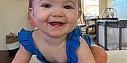 Paisley is registered to the contest to win money with this photo: baby, child, smiling, crawling, blue_dress, indoor, living_room, table, chair, ceiling_fan, window, floor, person, cute, happy, tooth, hair_bow, skin, furniture, home