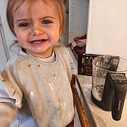 Athéna participe au concours pour gagner de l'argent avec cette photo : toddler, child, kitchen, flour, messy, smiling, bib, measuring_jug, countertop, wooden_chair, clothing, face, person, indoor, floor_tiles, cabinet, food_preparation, happy, young_child, playful