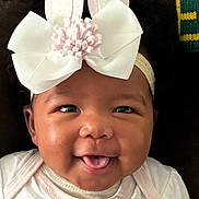 Parker is registered to the contest to win money with this photo: baby, child, face, headband, bow, bunny_ears, smile, tongue, cute, infant, closeup, portrait, happy, apparel, white_clothing, dark_background, expression, adorable, young, person