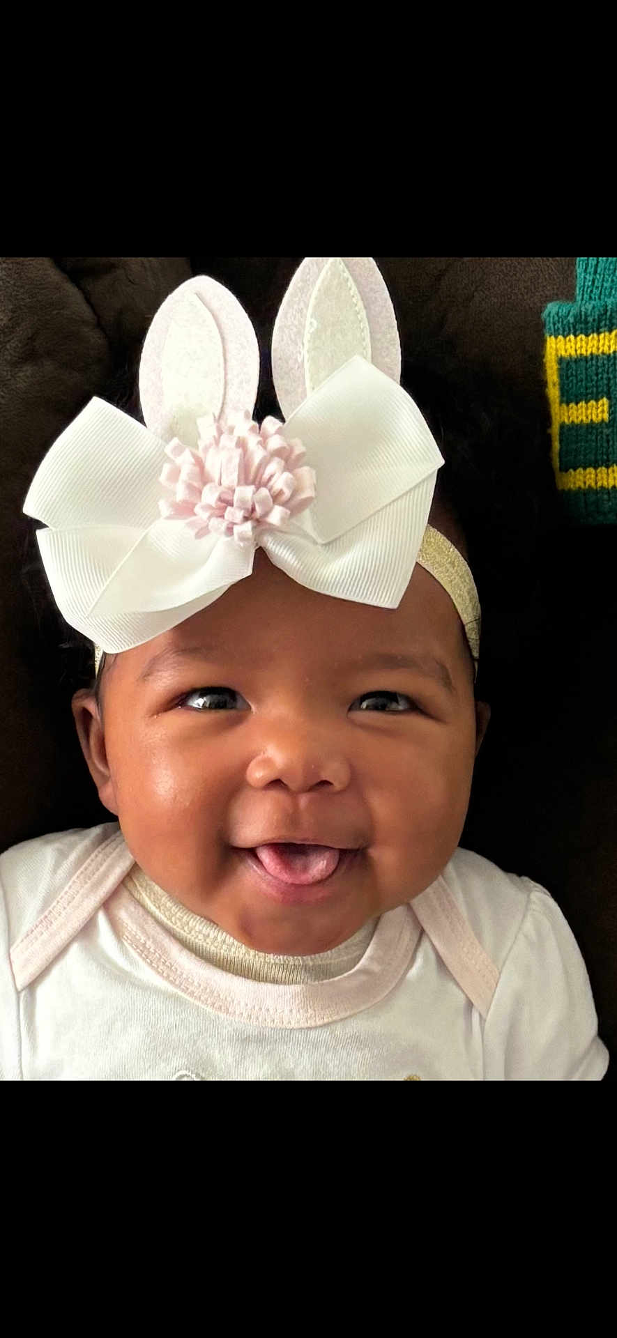 Parker is registered to the contest to win money with this photo: baby, child, face, headband, bow, bunny_ears, smile, tongue, cute, infant, closeup, portrait, happy, apparel, white_clothing, dark_background, expression, adorable, young, person