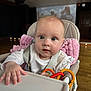 Verity is registered to the contest to win money with this photo: baby, high_chair, pink_blanket, toy_rings, curious, indoor, wooden_floor, child, infant, face, person, white_clothing, hand, seat_belt, tray, head, expression, cute, portrait, room