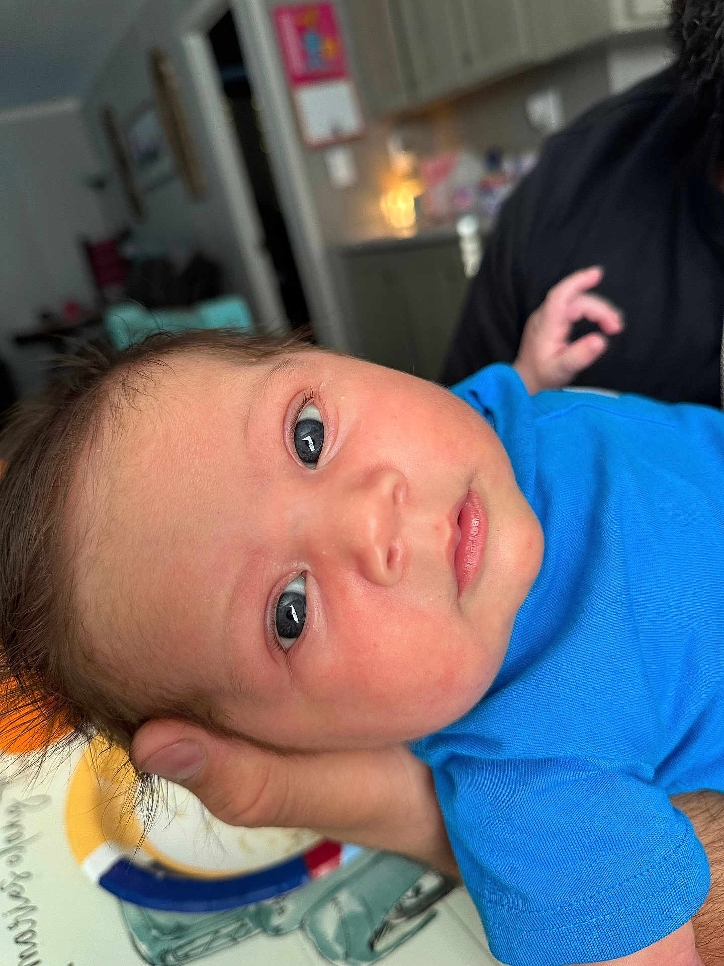 Jaxen joined the competition — help win amazing prizes! baby, blue_shirt, face, eyes, hand, indoor, kitchen, person, portrait, skin, table, plate, blurred_background, child, infant, resting, holding, home, casual, closeup