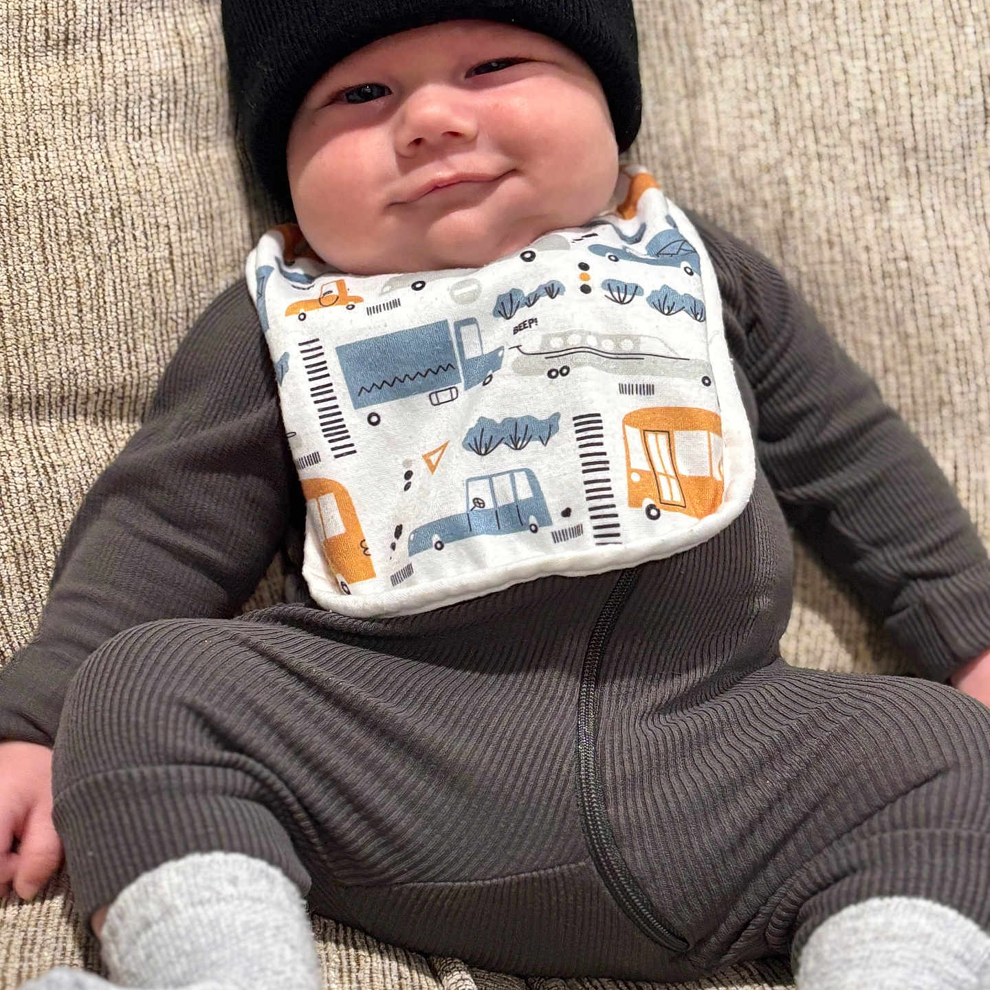 Tulsen joined the competition — help win amazing prizes! baby, beanie, bib, carhartt, couch, cozy, cute, face, footwear, gray_socks, indoors, infant, infant_clothing, lying_down, person, ribbed_fabric, smile, texture, young_child, zipped_onesie