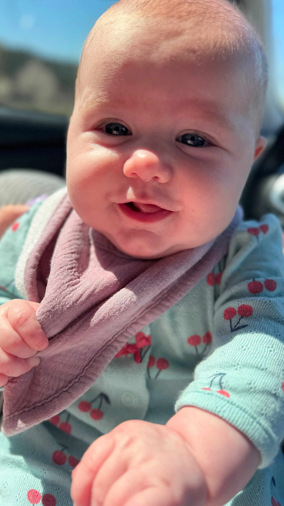 Haven joined the competition — help win amazing prizes! baby, infant, child, face, close_up, smiling, bib, onesie, cherry_pattern, hand, eyes, cheeks, forehead, eyelashes, sunlight, portrait, adorable, cozy, car_interior, shallow_depth_of_field