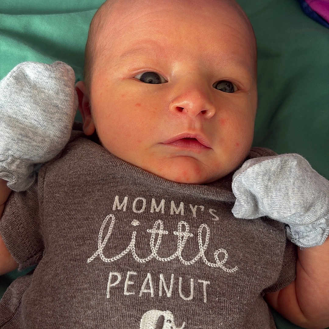 Klyde is registered to the contest to win money with this photo: baby, infant, newborn, onesie, mittens, lying_down, green_blanket, clothing, face, head, cute, person, portrait, small, child, skin, expression, indoors, text_on_clothing, elephant