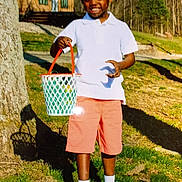 Jordan joined the competition — help win amazing prizes! child, boy, smiling, white_polo, salmon_shorts, basket, outdoor, grass, tree, wooden_cabin, sunlight, shoes, socks, nature, happy, daylight, portrait, playful, summer, casual_clothing
