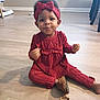 child, toddler, headband, bow, red_clothing, jumpsuit, sitting, wooden_floor, leopard_print_shoes, shoe, barefoot, smiling, playful, indoor, portrait, hands, eyes, hair, power_outlet, wall