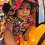 toddler, child, hat, colorful_clothing, yellow, play_vehicle, indoor, shirt, smiling, person, face, cartoon_pattern, bucket_hat, arm, play_area, restaurant, fun, kids, cute, background