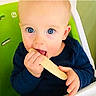 baby, child, infant, face, eyes, blue_eyes, smiling, eating, snack, food, hands, high_chair, indoor, portrait, sitting, navy_clothing, green_background, close_up, adorable, photography
